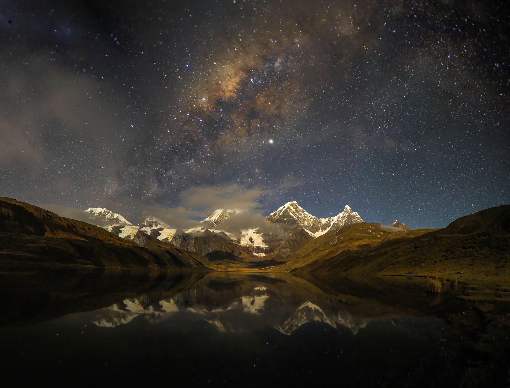 Snow-covered mountains under a starry night sky reflected in a lake