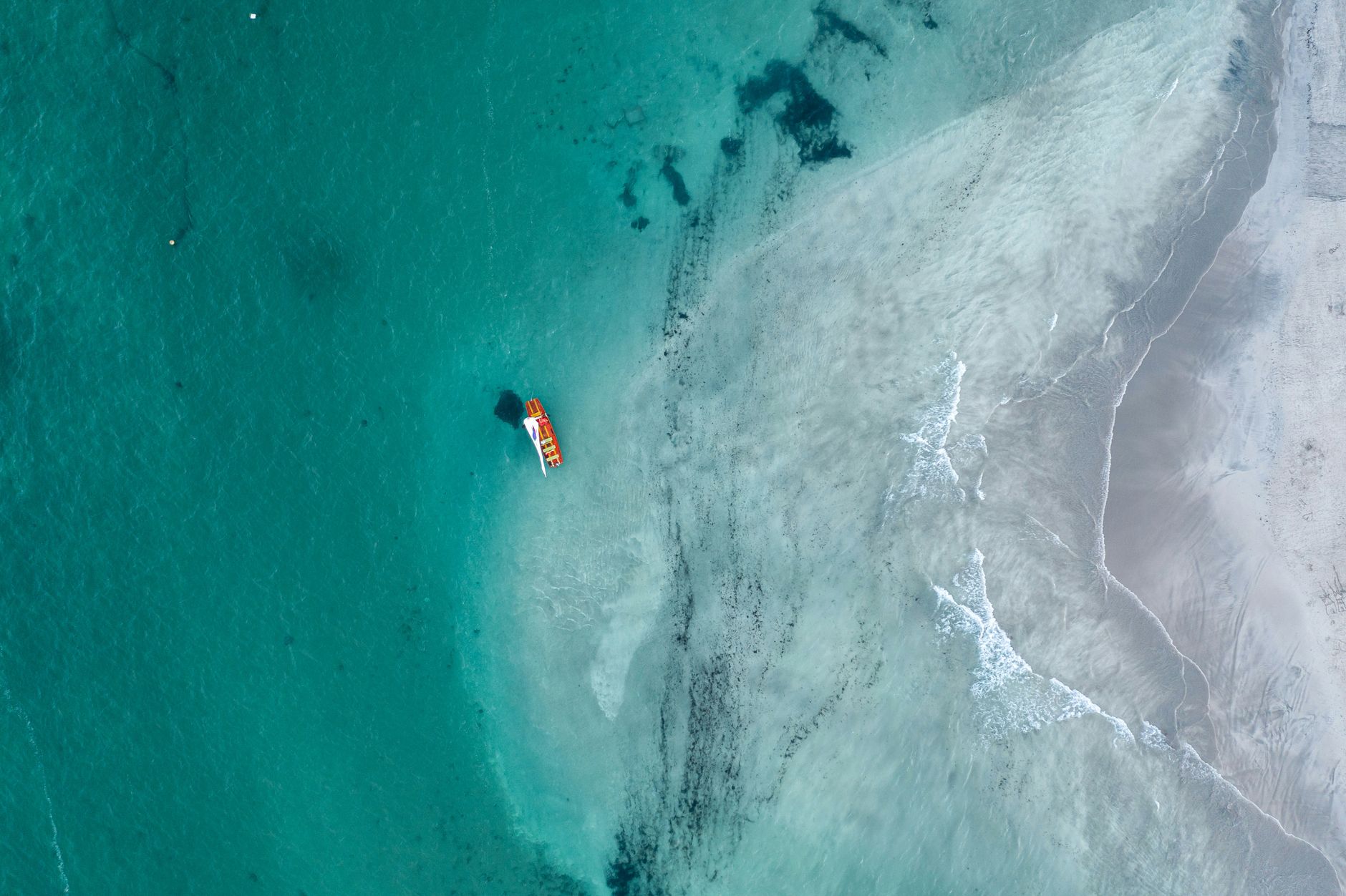 Aerial view of a lone sailboat on clear turquoise water near a sandy coast